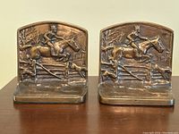 Pair of brass bookends with horse and rider jumping a fence with hunting dogs, front view.
