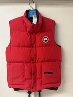 Front view of red sleeveless puffer vest hung on hanger showing full length with snap-button closure and patch pockets
