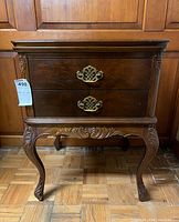 Front view of the carved wooden end table showing two drawers with metal handles and the detailed carvings on legs and apron.