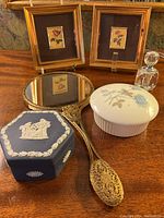 Overall view of the vanity items including two miniature framed floral prints, Wedgwood jasperware box, white ceramic box, cut glass perfume bottle, and ornate hand mirror.