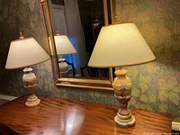 Pair of alabaster lamps on a wooden surface in front of floral wallpaper and a gold-framed mirror