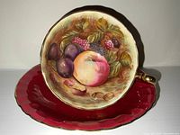 Front view of the teacup resting inside the red saucer showing detailed fruit painting and gold accents.