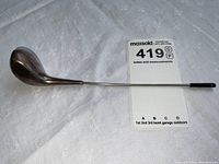 Full length view of a large sterling silver golf club styled object on a white surface with a MaxSold card showing lot number 419.
