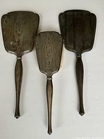 Backs of two hand mirrors and metal brush base without bristles, showing engraved decorative motifs and handle shapes.