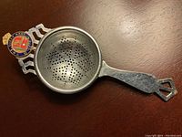 Top view of the silver tea strainer showing the perforated bowl with star-shaped hole pattern and the enamelled Queen Mary badge at the top end of the handle.