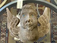 Front view of the carved angel head showing blue glass eyes and wing details, placed on a glass table with a patterned rug underneath.