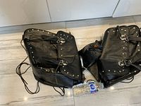 Pair of black leather motorcycle saddle bags placed on floor, showing overall look with silver studs and buckle straps, alongside a water bottle for scale.