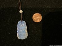 Close-up of translucent blue carved moonstone pendant on a brown cord next to a penny for scale