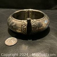 Photo of large vintage hinged silver-tone bangle with detailed etched and embossed floral patterns, closed and resting on black surface next to a quarter for size reference.