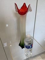 Full side view of the tall, mouth-blown glass vase beside a water bottle for size reference, showing the red ruffled rim and gradient coloring from white to green on the body with a flared base.