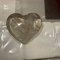 Photo of a large, polished natural quartz heart-shaped crystal resting on a white surface, showing its transparency and internal natural inclusions.