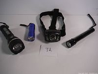 Four flashlights displayed on a table: large black flashlight, small blue flashlight, black headlamp with strap, and long black flashlight with hook.