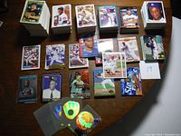 Photo shows multiple stacks of vintage baseball cards arranged on a wooden table, along with several loose cards displayed in front, featuring various players in batting and pitching stances, with varied card designs including color and black & white images.