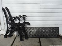 Two ornate cast iron bench side frames and one lattice metal backrest panel shown leaning against white siding on concrete floor.