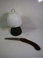 Coleman lantern and folding saw on a white surface, showing saw's wooden handle and folded metal blade.