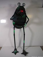 Full body view of tall black metal frog garden ornament with long legs and textured body showing green accents and red tongue detail.