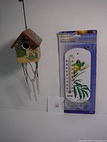 Full view of the packaged thermometer with frog design next to the hanging decorative birdhouse wind chime.