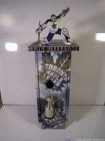 Front view of wooden birdhouse painted with Toronto Maple Leafs logo, miniature silver Stanley Cup below entrance hole, and goalie figurine on roof.