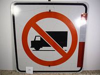 Front view of vintage no trucks traffic sign with black truck silhouette and red circular prohibition symbol on white background, showing mounting holes and wear.