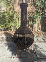 Full front view of rust-colored metal chimenea placed on outdoor stone patio, surrounded by garden area.