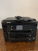 Front view of black Epson WF-3540 all-in-one printer on wooden surface, showing control panel and paper trays.