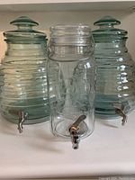 Front view of all three glass beverage dispensers showing two green tint bee hive style and one clear canning jar style with metal spigots and lids.