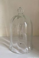 Front view of clear glass cloche with scalloped edge and rounded knob handle on top, placed on white surface with plain background.