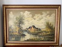 Full view of framed painting showing European-style cottage with trees and water, gold tone frame.