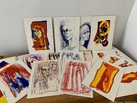 Wide view of 21 original pastel artworks arranged on a wooden surface, featuring abstract and figurative subjects with vibrant colors and varied media (watercolour paper, standard paper, board).