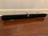 Front view of black Vizio sound bar placed on wooden floor, showing full length and rectangular shape.