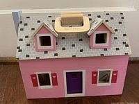 Front view of pink wooden doll house with gray tiled roof, purple front door, and handle on top.