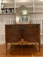 Front view of wood dresser showing three drawers with ornately veneered fronts and turned legs, topped with attached arched mirror.