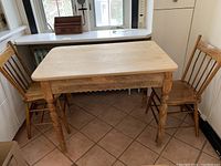View of farmhouse table with unfinished wood top and three wooden spindle-back chairs arranged around it.