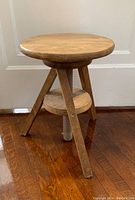 Side view of the wood stool showing the round seat, three legs, and central screw mechanism for height adjustment.