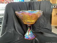 Front view of the amber carnival glass pedestal compote bowl on a black background showing grape clusters and leaf pattern with scalloped rim.