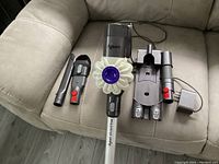Dyson vacuum and accessories displayed on light brown couch, showing main unit, crevice tool, combination brush tool, wall mount, charger, and wand.