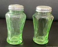 Front view of two green uranium glass salt and pepper shakers with metal lids, showing faceted hexagonal glass bodies and rounded bases.