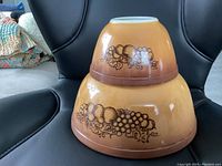 Two Pyrex bowls stacked, showing the distinct Old Orchard pattern with fruit and foliage in brown on an amber background fading darker at the base.