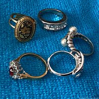 Top angle showing five rings arranged on blue fabric, highlighting the damascene oval floral ring, red stone ring, pearl ring, and two silver-tone rings.