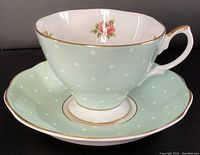 Side view of the mint green teacup with white polka dots, gold rim, and pink rose inside, sitting on matching saucer.