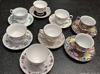 Full lot view showing seven teacups with matching saucers in varied floral patterns and colors on a black surface.