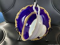 Photo showing white porcelain teacup with gold rim and handle detail, hanging on matching deep cobalt blue saucer with ornate gold trim on edges and central gold band.