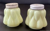 Pair of Victorian custard yellow opalware salt and pepper shakers with metal lids on black background.