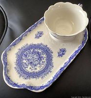 Porcelain teacup sitting on matching elongated dessert saucer showing blue floral decoration on off-white base with scalloped edges.