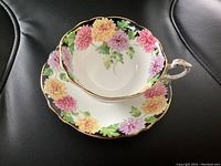 Top and side view of the fine porcelain Paragon teacup resting on the matching saucer. Both have a colorful chrysanthemum floral pattern and gold edging.