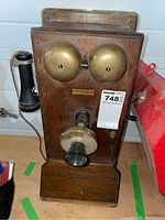 Front view showing oak backboard, dual brass bells, brass transmitter and black mouthpiece, earpiece on side hook, crank handle visible