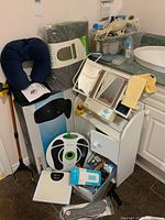 Wide view of lot showing foot massager device, neck pillow, carry bag, makeup mirror on storage cabinet, pill containers, plastic box with supplies, and more.