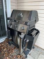 Full view of Broil King stainless steel gas BBQ grill with three knobs and folding side shelves and propane tank.