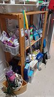 Wooden 3-tier shelf and contents including cleaning and household items, cords, buckets and light bulbs seen overall