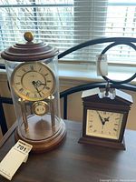 Photo showing both the Howard Miller anniversary clock and Jerger wind-up alarm clock side by side on a table near a window.